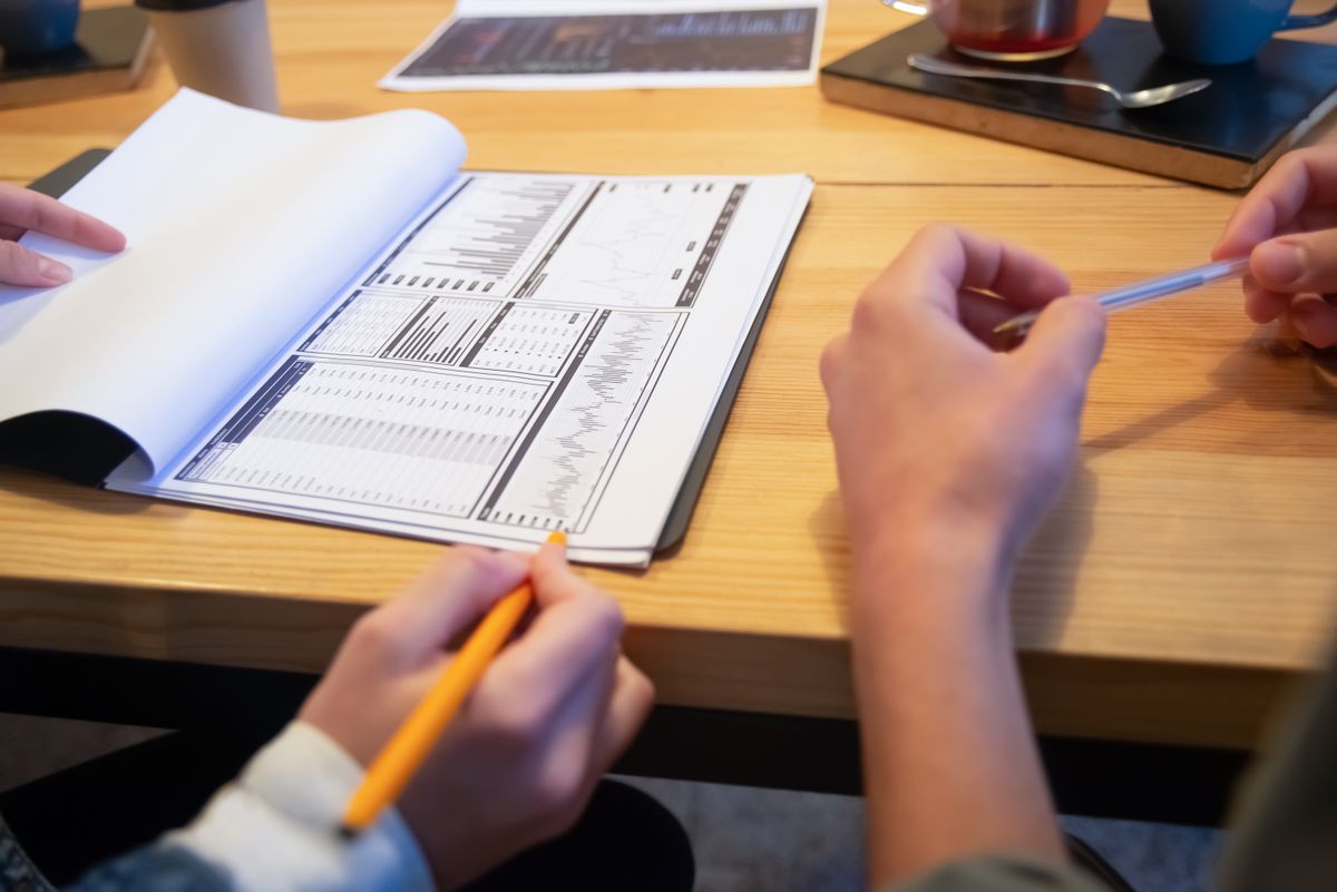 Two people with pens working over printed tables and charts on a wooden table