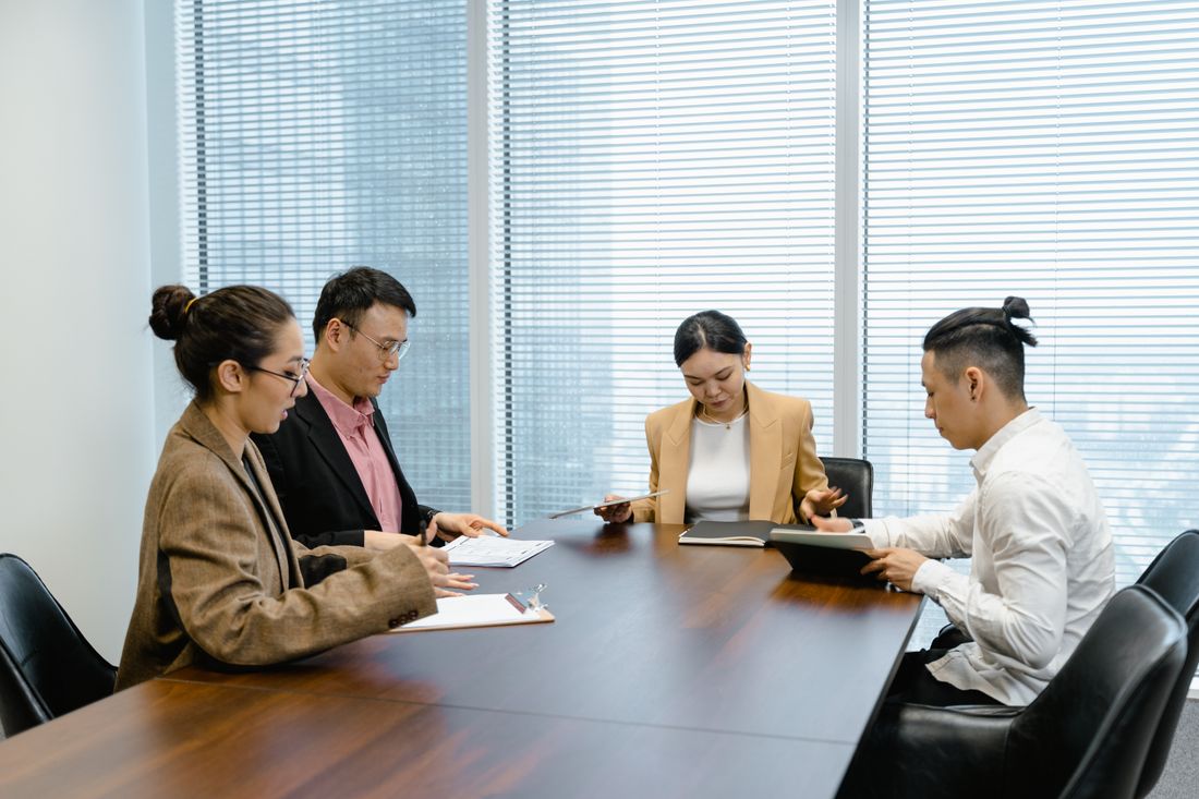 Four people in a meeting around a dark conference table, reviewing documents together by a bright window