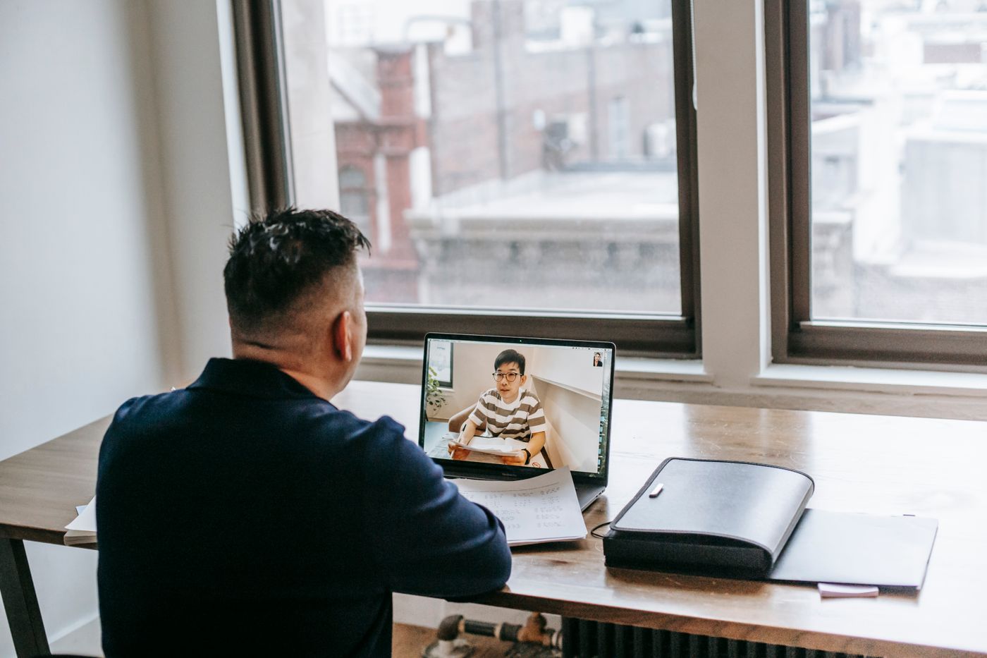 A video call in progress, seen over one person's shoulder: a laptop on a wooden desk by a large window, the other party listening on screen