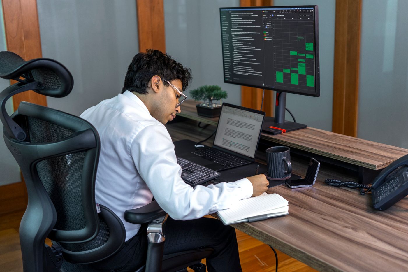 A person working at a desk between two live screens: notes on the laptop in front, the new system board running on the monitor beside it